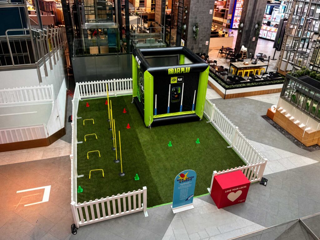 AFL Play custom inflatable activation cube installed with artificial turf and fencing inside a shopping centre concourse.