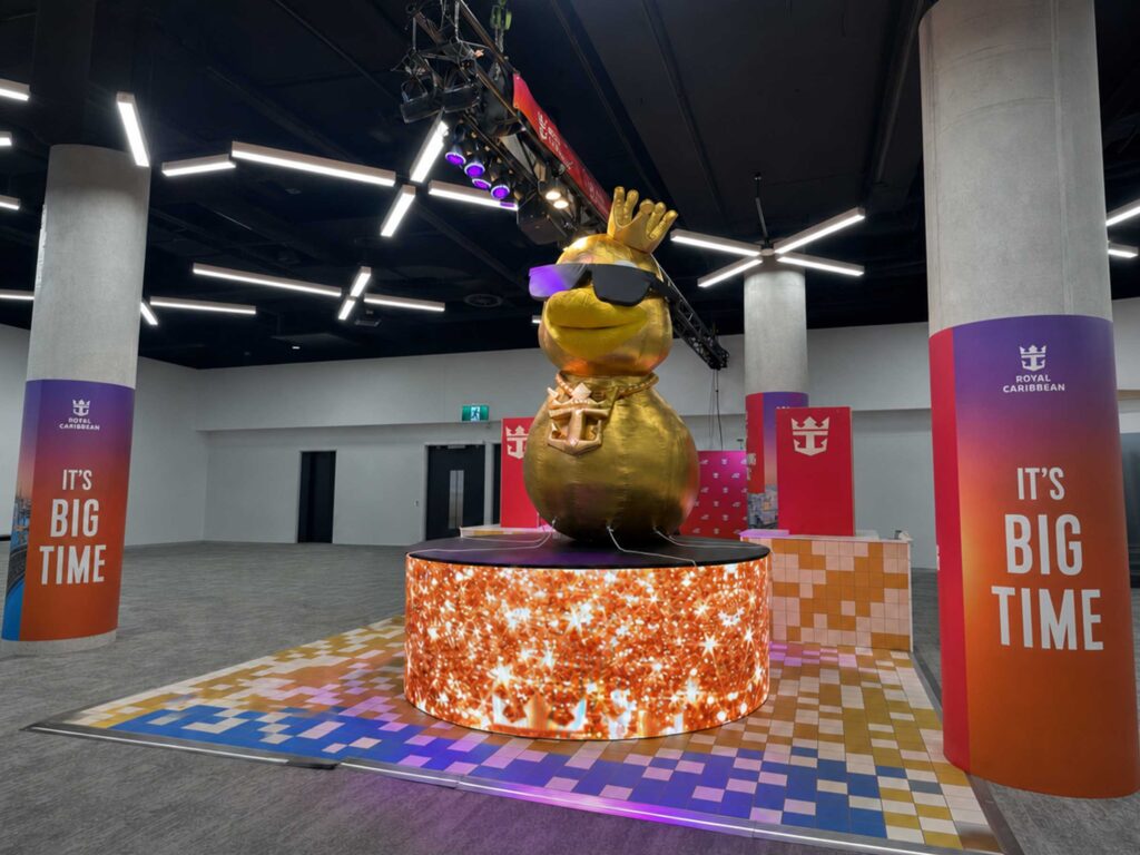 Custom inflatable replica duck wearing sunglasses, a crown, and an anchor chain on a digital plinth for Royal Caribbean Cruises trade show booth.